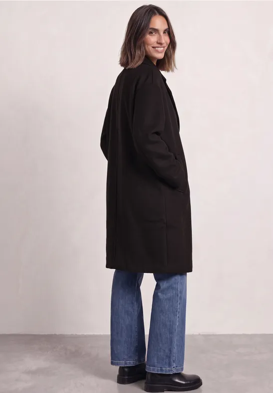 Eine Frau in schwarzem Mantel, blauen Jeans und schwarzen Schuhen steht seitlich vor hellem Hintergrund.