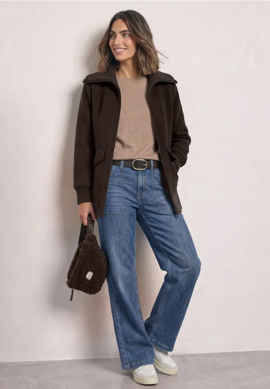 Frau in brauner Jacke, beigem Pullover, blauen Jeans und weißen Sneakern, hält eine braune Tasche.