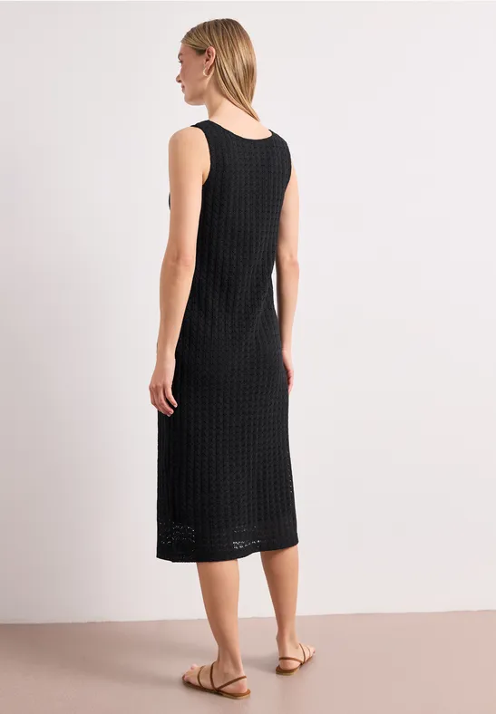 Frau im ärmellosen, schwarzen Midikleid mit Struktur, seitlich stehend, flache Sandalen, heller Hintergrund.