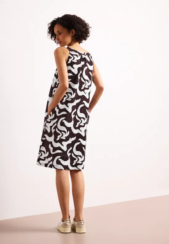 Frau von hinten im schwarz-weiß gemusterten, ärmellosen Midikleid mit Taschen und beigen Sneakern.