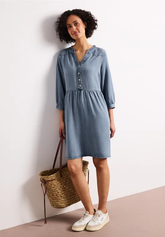 Vrouw in een casual denimjurk met knopen, witte sneakers en een gevlochten rieten tas