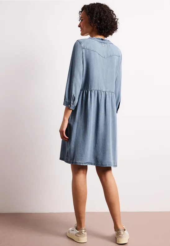 Vrouw van achteren in losvallende, knielange spijkerjurk met driekwartmouwen en sneakers tegen lichte achtergrond.