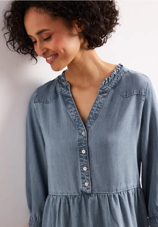 Vrouw draagt losvallende lichtblauwe denimjurk met ruches bij de kraag en knoopsluiting.