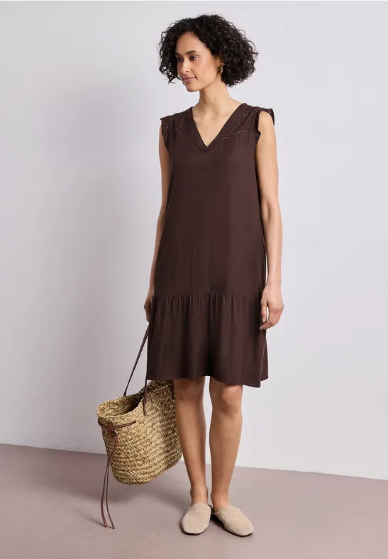 Femme en robe marron sans manches à volant, panier en paille et mules beiges devant fond neutre.