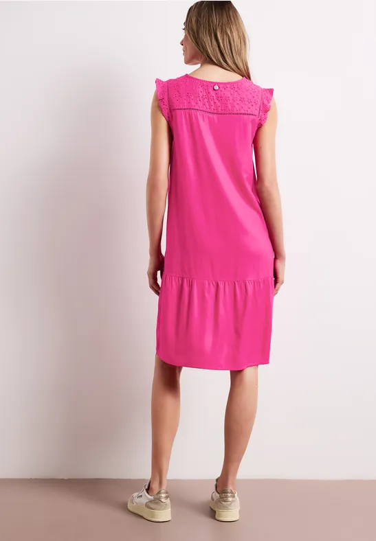 Vrouw in los vallende roze jurk tot op de knieën met kanten rugdetail en beige sneakers.