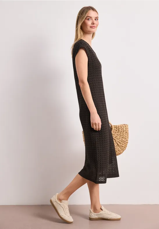 Femme en robe midi noire texturée, baskets claires et panier en osier sur fond blanc.