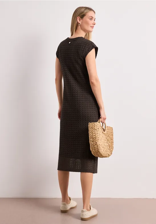 Femme en robe mi-longue noire à motif discret, manches courtes, sac en paille et chaussures à semelle épaisse.
