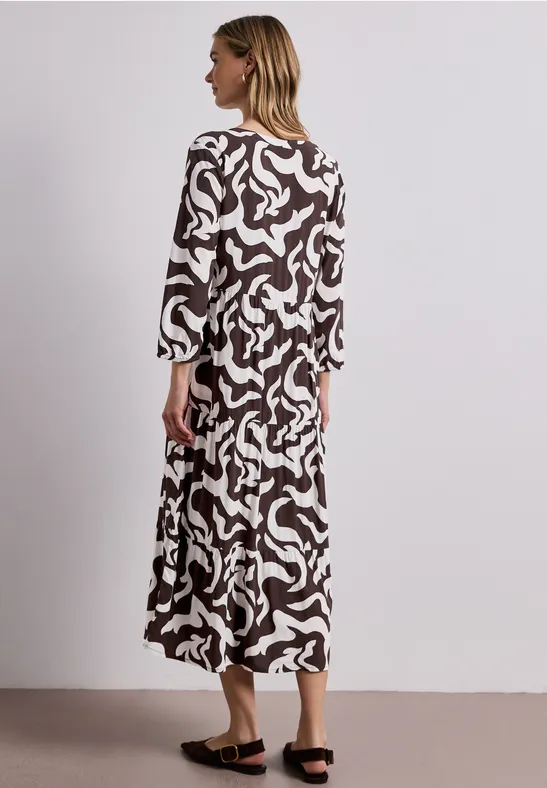 Frau im langen, fließenden Kleid mit braun-weißem abstraktem Muster vor neutralem Hintergrund.