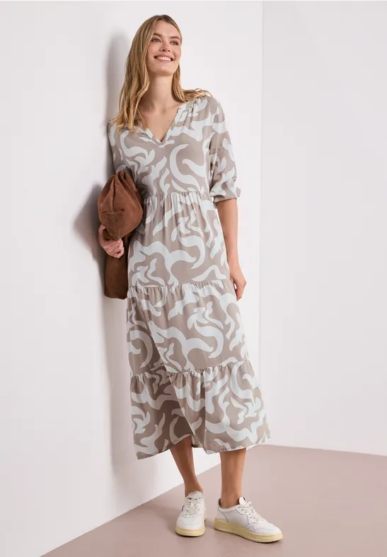 Een vrouw in een luchtige, gebloemde midi-jurk leunend tegen een witte muur, met witte sneakers en clutch.