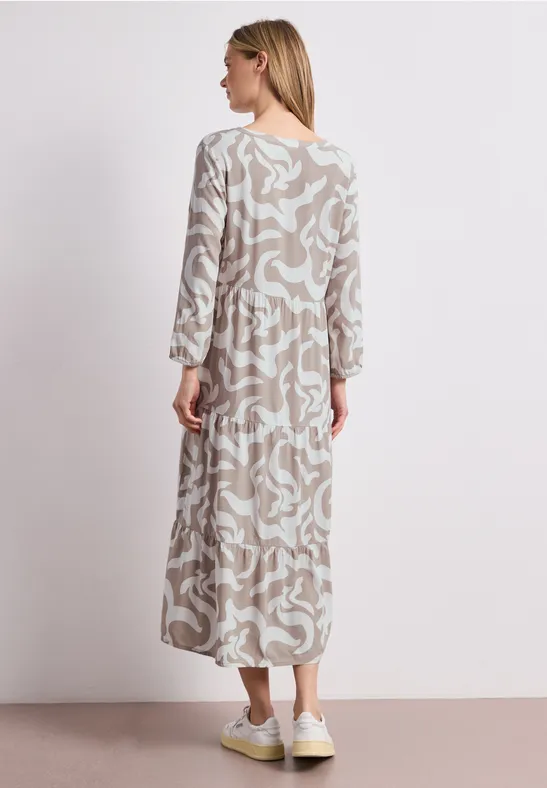 Een vrouw van achteren in een beige-witte maxi-jurk en witte sneakers voor een lichte achtergrond.