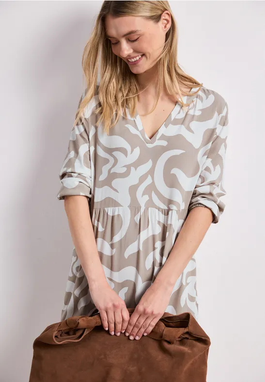 Vrouw in beige midi-jurk met print, V-hals en driekwartmouwen, met een bruine leren tas in de hand.