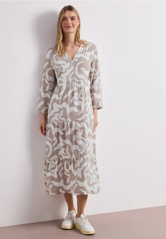 Vrouw in een beige midijurk met wit abstract dessin, V-hals, gecombineerd met witte sneakers