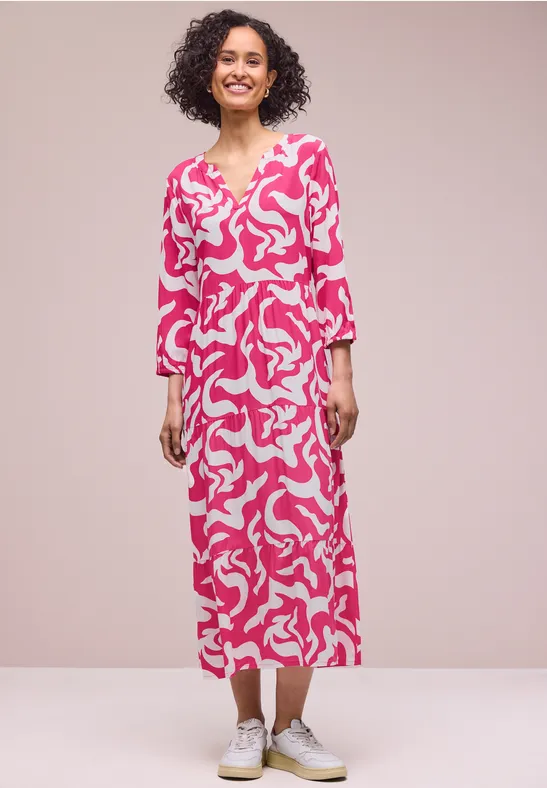 Frau in einem knöchellangen pink-weißen Kleid mit abstraktem Muster und weißen Sneakern.