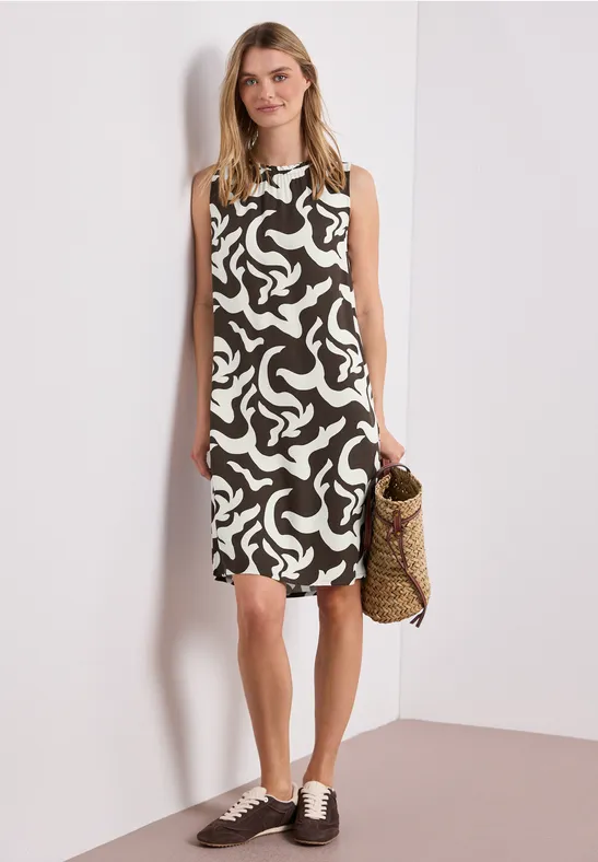 Vrouw in zwart-witte, mouwloze jurk met patroon, houdt strooien tas, draagt bruine veterschoenen