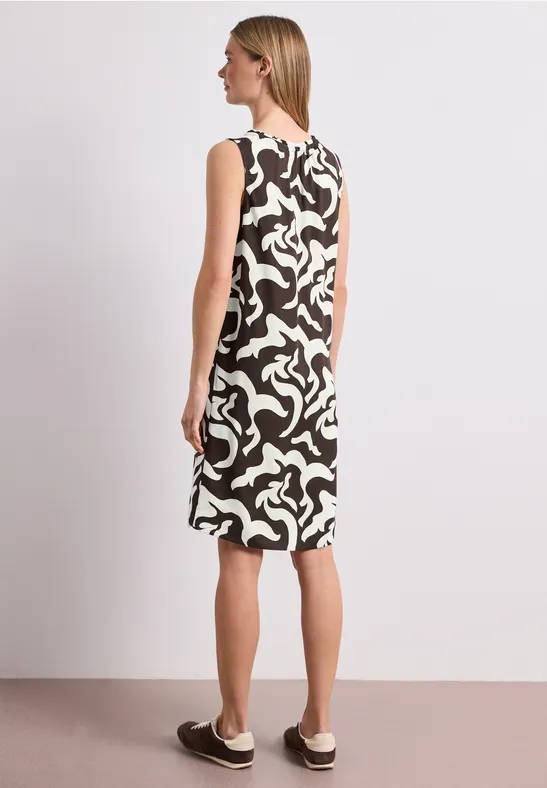 Rugaanzicht van een vrouw in een mouwloze zwart-witte jurk met abstract patroon en platte instapschoenen.