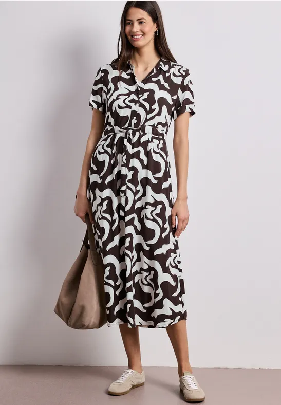 Frau in Midi-Kleid mit abstraktem Schwarz-Weiß-Muster, V-Ausschnitt, weißen Sneakern und Tasche.