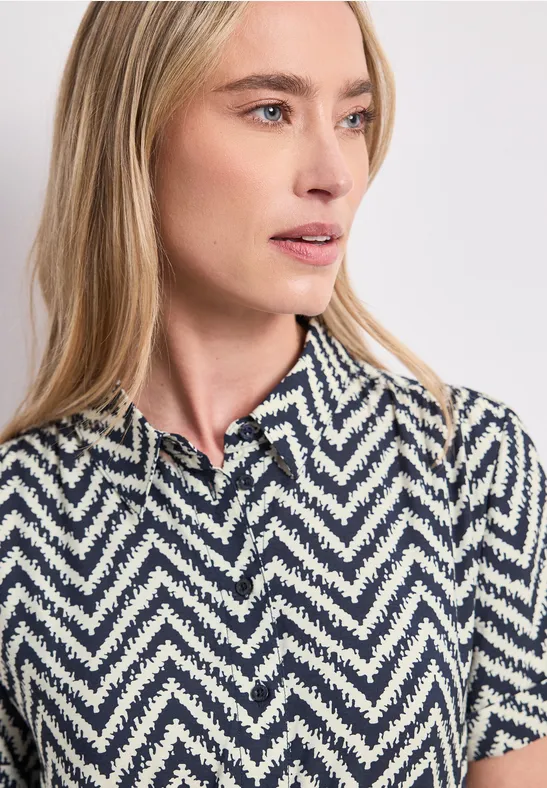 Blonde Frau trägt kurzärmliges, weiß-blaues Zickzack-Poloshirt mit Kragen.