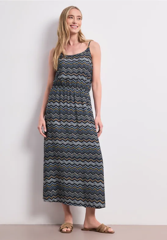 Frau in langem, ärmellosem Kleid mit buntem Zickzackmuster, Sandalen und weißem Hintergrund.