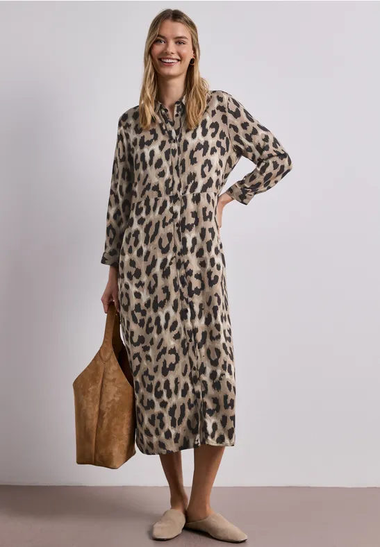 Vrouw in lange luipaardjurk met grote bruine tas en beige schoenen.