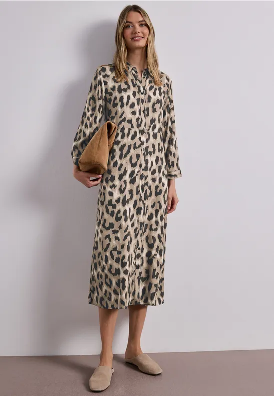 Vrouw in midi-jurk met luipaardprint en lange mouwen, beige schoenen en clutch, neutrale achtergrond.