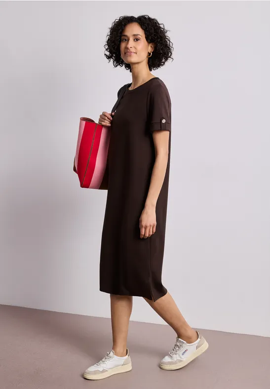 Frau im lockeren dunkelbraunen Midikleid trägt weiße Sneaker und eine rote Tasche vor schlichtem Hintergrund.