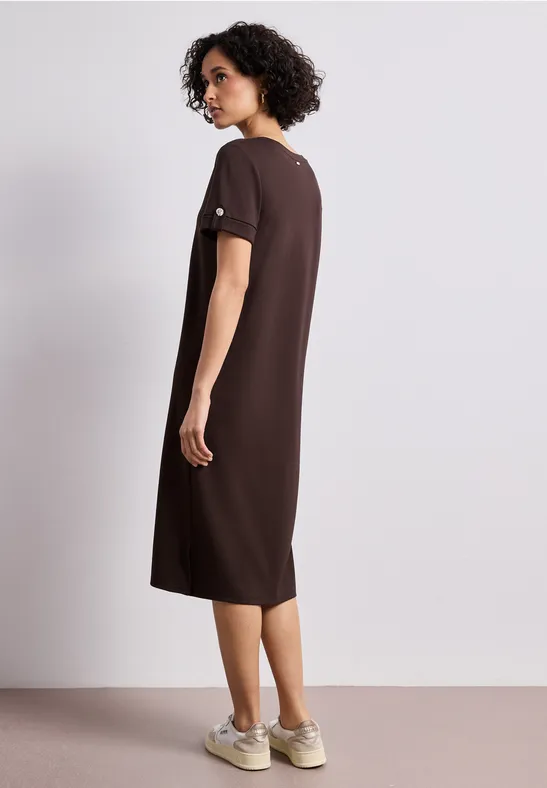 Frau von hinten in lockerem braunem Midi-Kleid und Sneakern, auf neutralem Hintergrund.