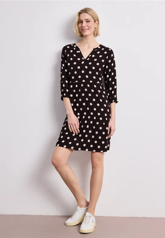 Frau in schwarzem Polka-Dot-Kleid mit 3/4-Ärmeln und weißen Sneakers, stehend vor weißem Hintergrund.