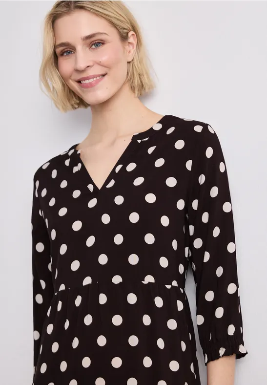 Frau in schwarzer Tupfenbluse mit V-Ausschnitt und 3/4-Ärmeln.