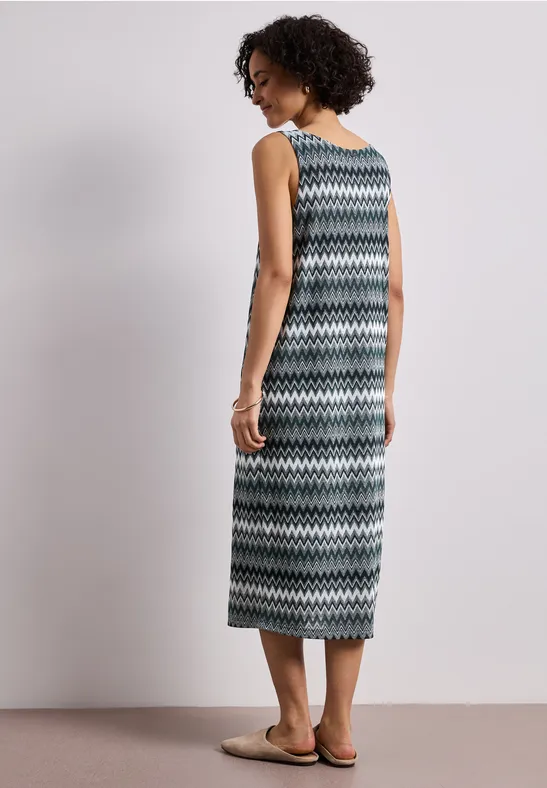 Frau im Maxikleid mit Zickzack-Muster, trägt Sandalen, steht vor neutralem Hintergrund.