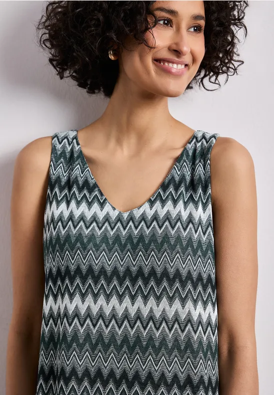 Person mit lockigem Haar trägt ärmelloses V-Ausschnitt-Top mit grauem, weißem und schwarzem Zickzackmuster.