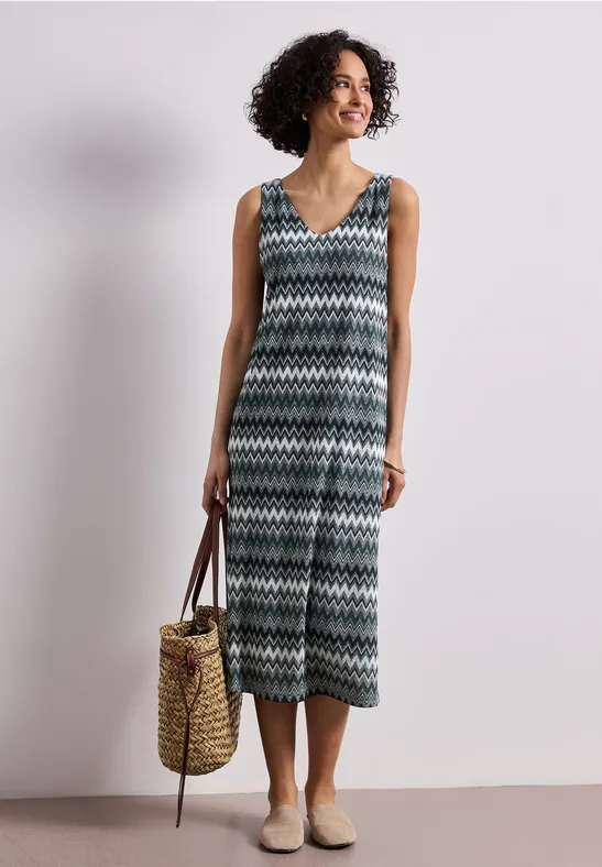 Frau trägt ärmellosen V-Ausschnitt-Jumpsuit mit schwarz-weiß-grünem Zickzackmuster, Strohtasche und beigen Schuhen.