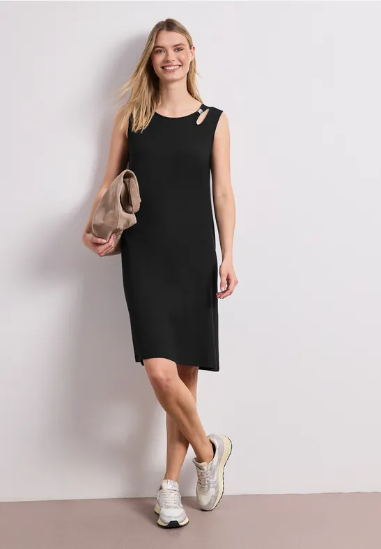 Frau in ärmellosem schwarzen Kleid mit Clutch und Turnschuhen vor hellem Hintergrund.