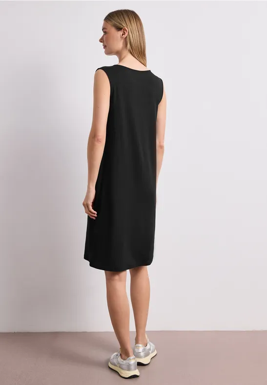 Frau von hinten in ärmellosem schwarzem knielangem Kleid und weißen Sneakern vor hellem Hintergrund.