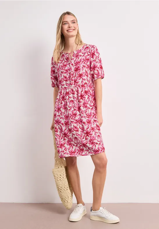 Femme en robe fluide rose à fleurs blanches, baskets blanches et sac en paille