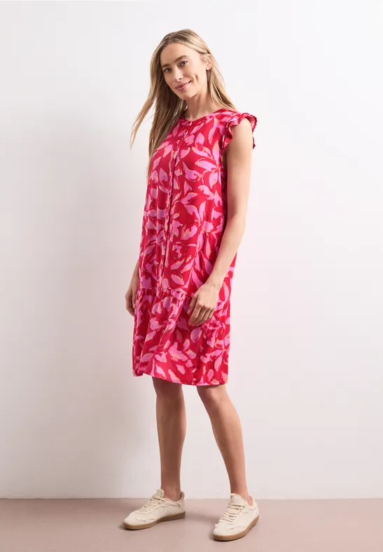 Femme portant une robe rose à motifs blancs jusqu’aux genoux et des baskets claires sur fond blanc.