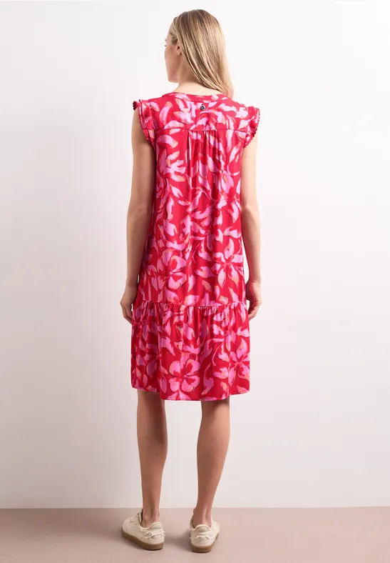Femme de dos portant une robe rouge à fleurs blanches jusqu’aux genoux et des chaussures plates.