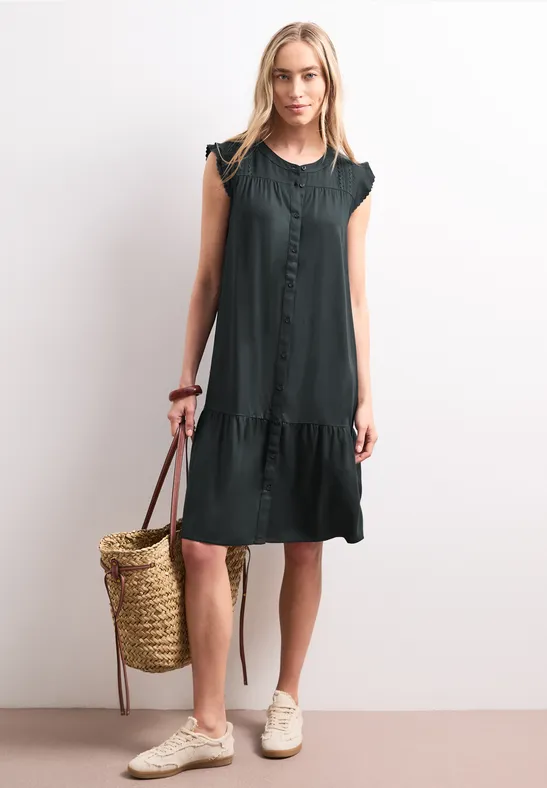 Femme porte une robe sans manches foncée jusqu'aux genoux, tient un sac en paille devant un fond clair.