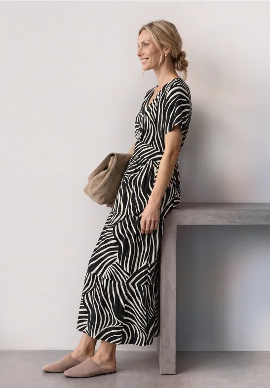 Femme en robe midi zébrée, tenue décontractée avec sac, appuyée sur une table devant un mur clair.