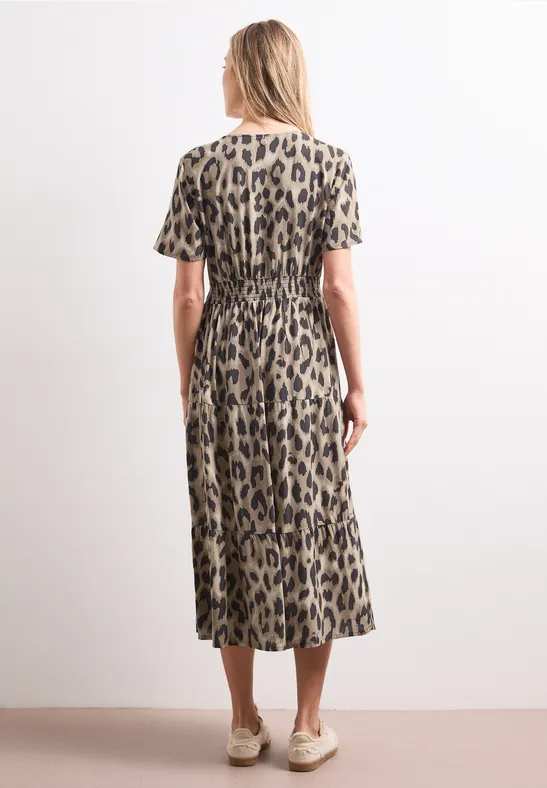 Rückansicht einer Frau in Midi-Kleid mit Leopardenmuster, kurzen Ärmeln und beigen Schuhen vor weißer Wand.
