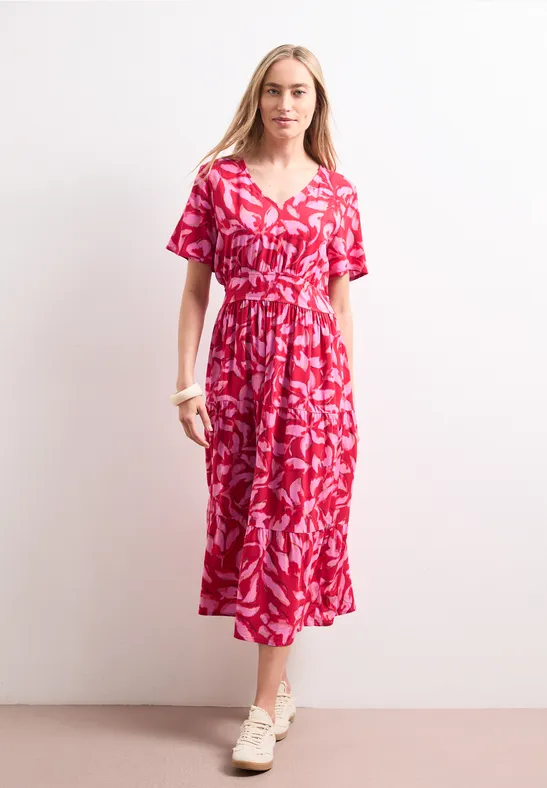 Une femme porte une robe midi rouge imprimée et pose devant un mur clair minimaliste.