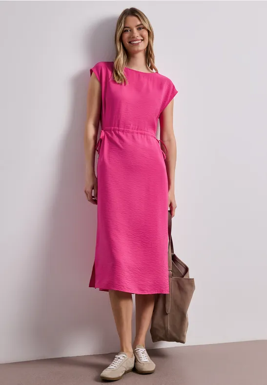 Eine Frau trägt ein pinkes Midi-Kleid, weiße Sneaker und hält eine braune Tasche.