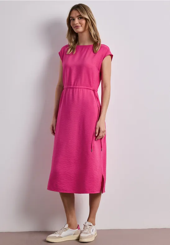 Frau in knielangem pinken Kleid und weißen Sneakers vor hellem Hintergrund.