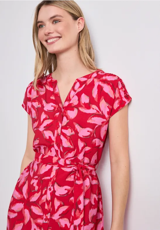 Vrouw draagt rode jurk met korte mouwen en roze bladprint, V-hals en tailleceintuur.
