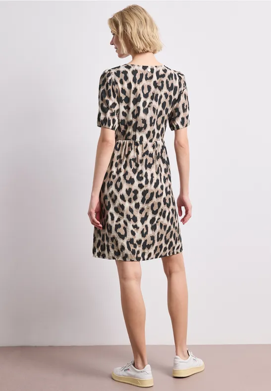 Frau von hinten in kurzem, leo-getupftem Kleid mit kurzen Ärmeln und hellen Sneakern vor neutralem Hintergrund.