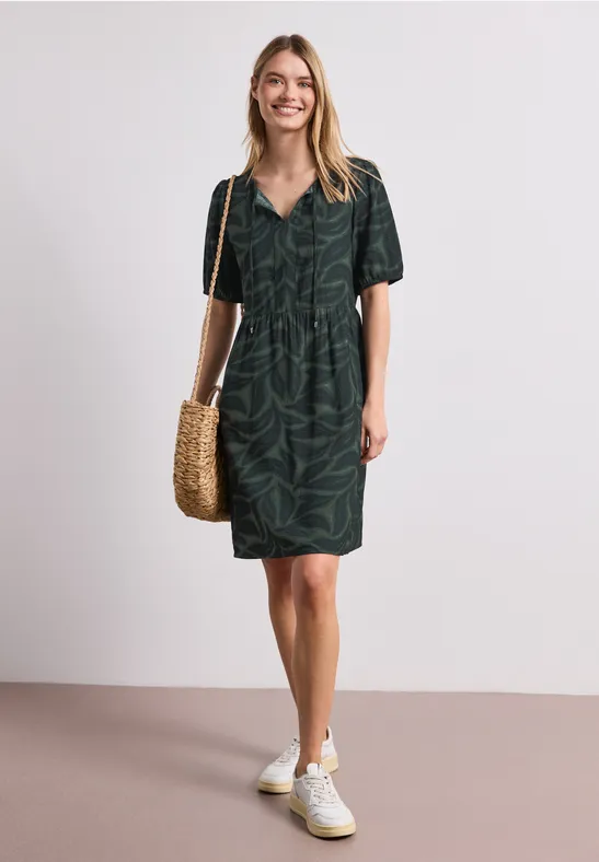 Vrouw in donker groen overhemdjurk met bladprint, witte sneakers en gevlochten strooien tas.