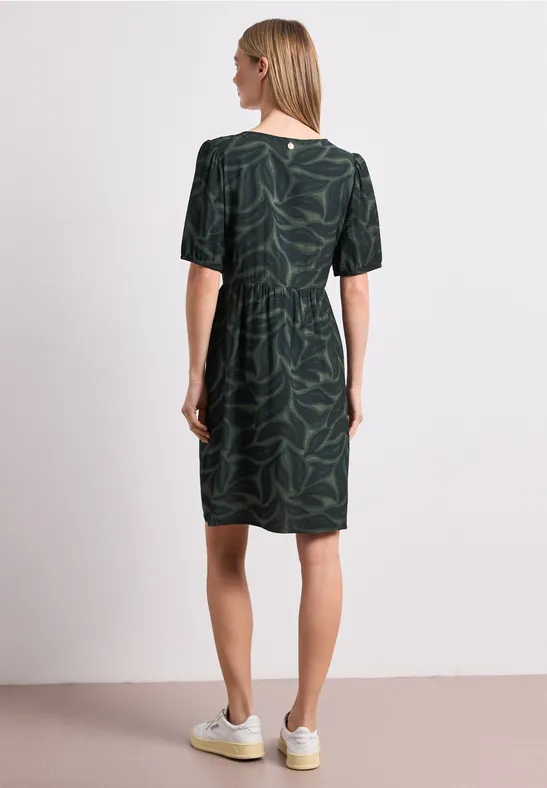 Vrouw van achteren in groene jurk met bladprint en witte sneakers voor witte achtergrond.