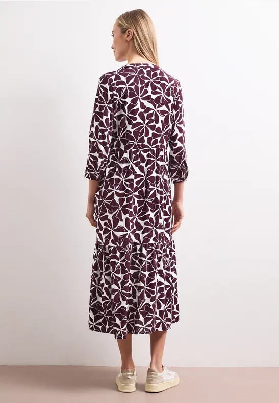 Rückansicht einer Frau im knöchellangen, violetten Kleid mit weißem Blattmuster und hellen Sneakern.