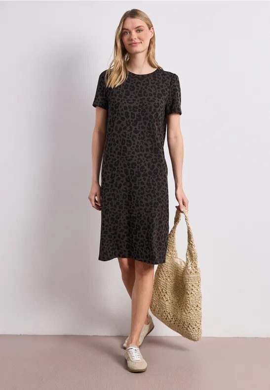 Vrouw in zwart gemusterd kortarmjurk met strooien tas en witte instappers.