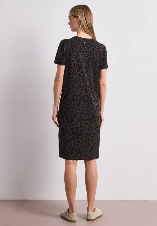 Achteraanzicht van een vrouw in een knielange zwarte jurk met subtiel patroon, korte mouwen, blond haar, voor een neutrale achtergrond.