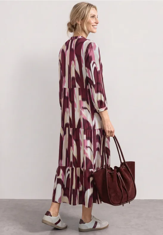 Vrouw van achteren in een losvallende maxi-jurk met abstract motief, met een bordeaux tas en sandalen.